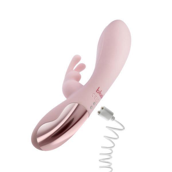 The Morgan Beginner G-Spot Rabbit Vibrator by Blush is a blush pink silicone sex toy with a sleek, curved design that features a clitoral stimulator attachment and a metallic accent towards the base. The device comes with a coiled white charging cable and offers intense clitoral stimulation.