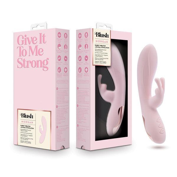 The image shows a pink personal massager made of silky silicone and its box packaging from the brand Blush. The box displays the product name "Morgan Beginner G-Spot Rabbit Vibrator" and has light pink and white design elements, including text that reads "Give It To Me Strong.