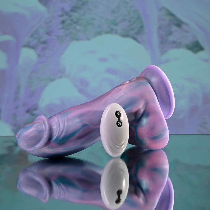 The Mythical Long Thick Vibrating Fantasy Dildo in Pastel Ombre, featuring swirled purple, blue, and pink silicone, rests on a reflective surface next to its white wireless remote against a blurred abstract background.