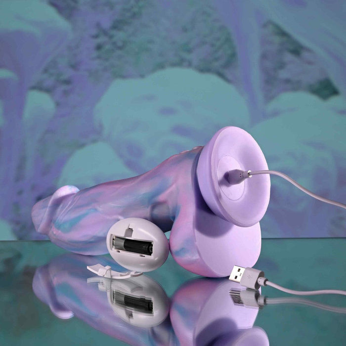 The Mythical Long Thick Vibrating Fantasy Dildo in Pastel Ombre, a silicone toy with a suction cup base and strap-on harness compatibility, rests on a reflective surface, charging via USB with its battery compartment cover removed.