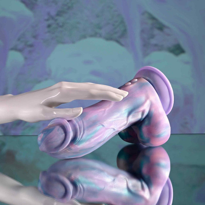 The Mythical Long Thick Vibrating Fantasy Dildo in Pastel Ombre, with swirls of purple, blue, and pink silicone, stands on a reflective surface as a pale hand gently touches the harness-compatible toy. The background echoes its matching pastel hues.