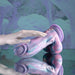 The Mythical Long Thick Vibrating Fantasy Dildo in Pastel Ombre, with swirls of purple, blue, and pink silicone, stands on a reflective surface as a pale hand gently touches the harness-compatible toy. The background echoes its matching pastel hues.