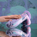 A manicured hand rests on the Mythical Long Thick Vibrating Fantasy Dildo in Pastel Ombre, made of body-safe silicone, showcased on a reflective surface with a soft, abstract background.