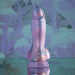 The Mythical Long Thick Vibrating Fantasy Dildo in Pastel Ombre, made from body-safe silicone, stands upright on a reflective surface, set against a soft, abstract purple and blue background.