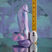 The Mythical Long Thick Vibrating Fantasy Dildo in Pastel Ombre stands upright beside a yellow measuring tape and a small white oval device with a question mark, all arranged on a reflective surface with a blurred background.