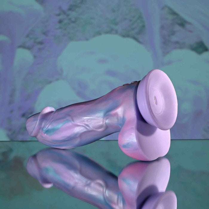 The Mythical Long Thick Vibrating Fantasy Dildo in Pastel Ombre rests on a reflective surface, set against a soft purple and blue abstract background.