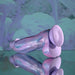 The Mythical Long Thick Vibrating Fantasy Dildo in Pastel Ombre rests on a reflective surface, set against a soft purple and blue abstract background.
