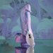 The Mythical Long Thick Vibrating Fantasy Dildo in Pastel Ombre stands upright on a round base, featuring a swirled pink, purple, and blue silicone design. A white oval remote with a power button is in front. The background is abstract and purple-toned.