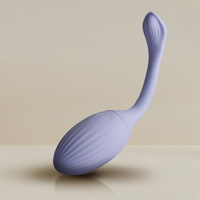 The Niya Rechargeable Kegel Exerciser and Vibrator with Remote is a light purple, elongated silicone massager with a rounded textured base and flexible curved handle, shown on a smooth surface against a neutral background.