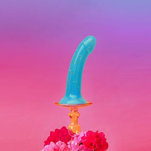 A Nightfall Teal Glitter 7 inch Silicone Dildo by Lovely Planet stands upright against a gradient background of pink and purple. The hands-free suction cup base sits among vibrant pink flowers, adding a colorful and artistic touch to the composition.