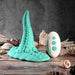 The Octopus Tentacle Insertable Remote Controlled Grinder in turquoise and its white remote are displayed on a wooden surface among seashells, set against a textured gray background.