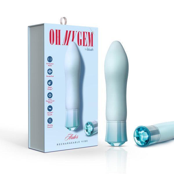 A white and light blue USB rechargeable vibrator stands next to its packaging, labeled "Oh My Gem Ardor Aquamarine Warming Internal & External Tapered Vibrator by Blush." The box features product highlights, a jeweled detail at the bottom matching the design of the vibrator, and information about its warming function.