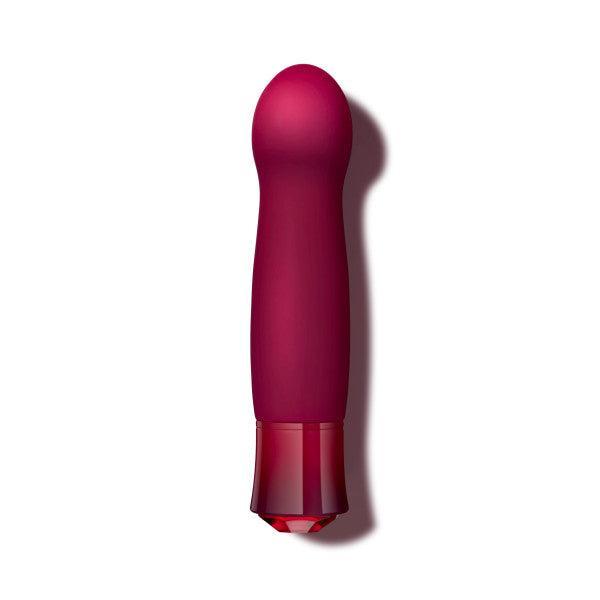 The Oh My Gem Classy Garnet Warming Internal & External Vibrator by Blush, a red, battery-operated vibrator with a smooth, slightly tapered shape and a rounded tip, stands upright against a plain white background. The vibrator features vibrating functions and has a darker red base that appears to serve as the control panel.