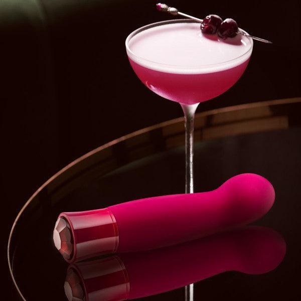 A vibrant pink cocktail in a martini glass garnished with cherries on a pick is displayed on a dark, reflective surface. Beside it lies the sleek Blush Oh My Gem Classy Garnet Warming Internal & External Vibrator, featuring vibrating functions and a garnet gemstone detail on the handle.