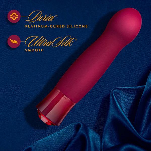 A red, sleek, curved silicone vibrator rests elegantly on a dark blue satin fabric. The surrounding text highlights its features, including "Luria™ Platinum-cured Silicone" and "UltraSilk® Smooth." Boasting vibrating functions and adorned with a gem-like garnet gemstone base, the Oh My Gem Classy Garnet Warming Internal & External Vibrator by Blush promises unparalleled pleasure with its elegant design.