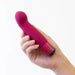 A hand holding the Blush Oh My Gem Classy Garnet Warming Internal & External pink, curved, silicone vibrator with multiple vibrating functions against a plain white background.