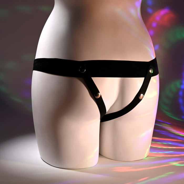 A mannequin torso showcases the Our First Hollow Strap-On Dildo + Harness Set - Vanilla, a minimal black harness-style lingerie piece compatible with a hollow strap-on, against a backdrop with colorful light reflections.
