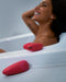 A woman smiling joyfully in a bathtub while a We-Vibe Chorus Remote & App Controlled Couples' Vibrator - Pink, featuring touch-sensitive vibrations, rests on the edge.
