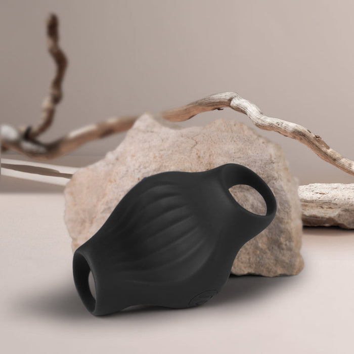 The Palm Vibrating Versatile & Accessible Masturbator, made of black body-safe silicone with a textured surface and dual openings, is displayed in front of a beige rock and dried wooden branches against a neutral background.