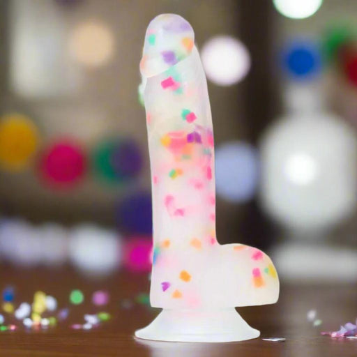 The Party Marty 7.5 Inch Silicone Rainbow Confetti Dildo stands upright on a wooden surface, showing off its translucent frosted silicone, embedded colorful confetti, and sturdy suction cup base against a festive backdrop.