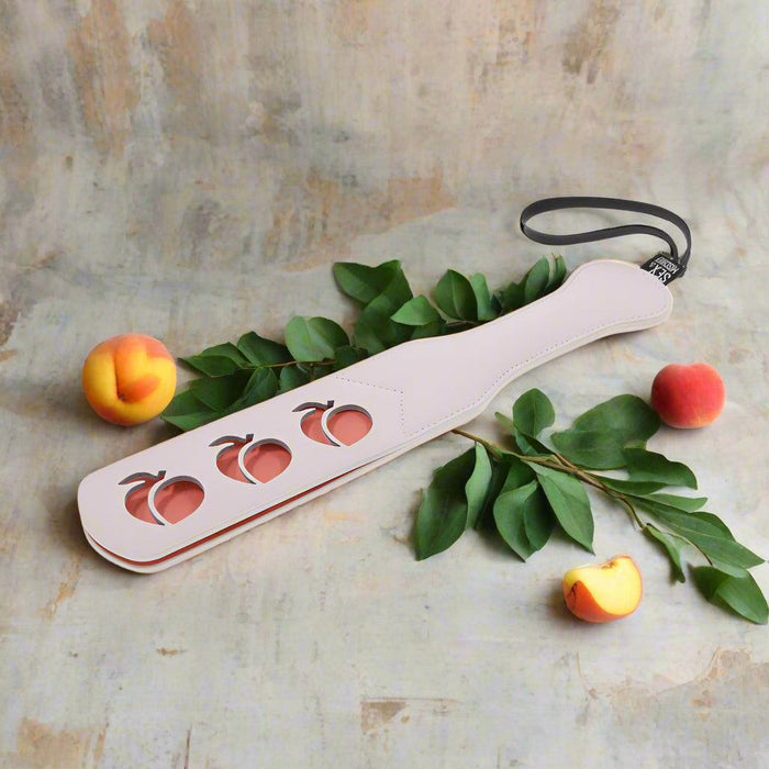 The Sportsheets Peaches 'n Cream Impression Paddle features a sleek white design with a black faux leather handle and a secure wrist strap. It includes three distinct peach-shaped cutouts, making it ideal for impact play enthusiasts who appreciate both style and function.