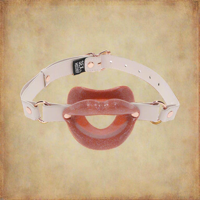 The Peaches 'n Cream Silicone Lips Open Mouth Gag by Sportsheets is featured against a white backdrop, showcasing its red, glittery design with white adjustable straps and metal buckles. This eye-catching accessory, shaped like lips, is ideal for those looking to add some sparkle to their kinky play.