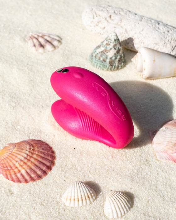 A vibrant pink We-Vibe Chorus Remote & App Controlled Couples' Vibrator nestled among seashells and coral on a sandy beach.