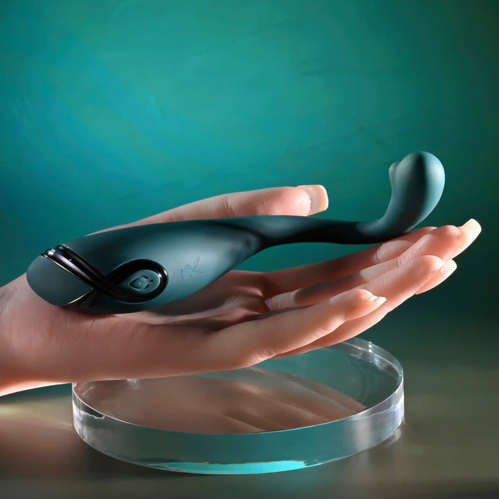 A realistic mannequin hand holds the Pinpoint Perfection Flexible Slim Vibrator, a sleek, curved, dark green body-safe silicone toy with buttons, displayed on a clear round stand against a teal background.