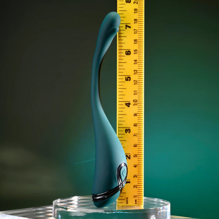 The Pinpoint Perfection Flexible Slim Vibrator, in teal, stands upright in a clear glass container next to a yellow measuring tape displaying its height against a green gradient background.
