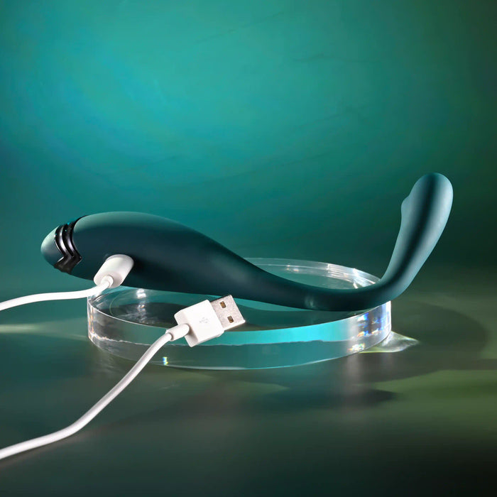 The Pinpoint Perfection Flexible Slim Vibrator, in dark green body-safe silicone, charges via a white USB cable while resting on a transparent round stand against a gradient green background.