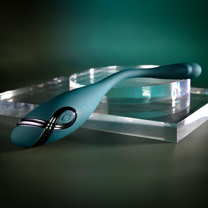 The Pinpoint Perfection Flexible Slim Vibrator, a sleek teal device with a metallic accent and curved design, is displayed on a clear rectangular acrylic platform against a dark green background.