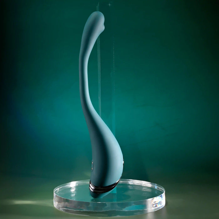 The Pinpoint Perfection Flexible Slim Vibrator, a sleek teal silicone device with a curved, smooth design, stands upright on a clear round base against a green gradient background.