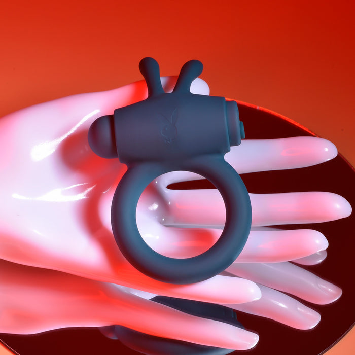 A Playboy Bunny Buzzer Vibrating Silicone Cock Ring in blue, with two small protrusions, rests on a white mannequin hand on a reflective surface lit by red and orange lights.