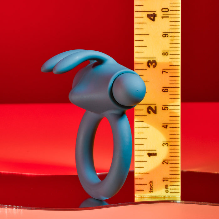 A Playboy Bunny Buzzer Vibrating Silicone Cock Ring in blue, made of body-safe silicone with rabbit ear extensions, stands upright by a yellow ruler (approx. 3 inches), set against a vivid red background.