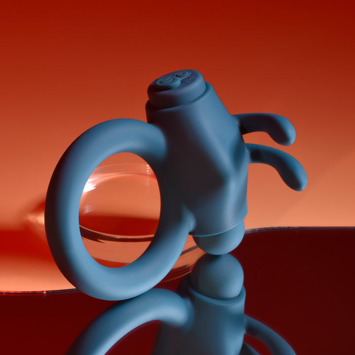 A Playboy Bunny Buzzer Vibrating Silicone Cock Ring in blue, featuring two flexible protrusions, is shown on a reflective surface with a red gradient background.