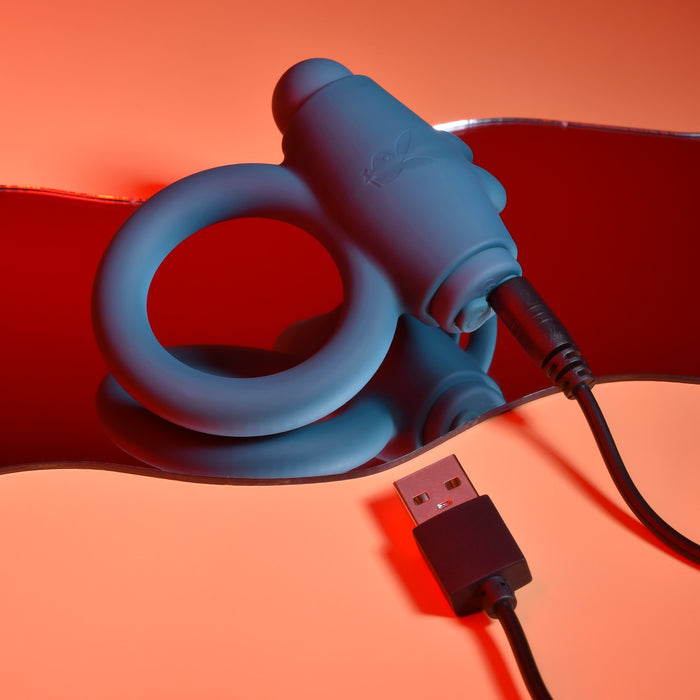 A Playboy Bunny Buzzer Vibrating Silicone Cock Ring in blue, made of body-safe silicone with a vibrating bullet, is plugged in and charging via USB on a reflective red and orange surface.