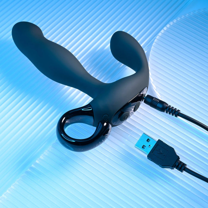 The Playboy Come Hither Motion Vibrating Prostate Massager by Evolved Novelties is showcased on a glossy, textured blue surface. The black device features a sleek, modern design with a loop handle and a curved, dual-ended structure that offers 10 vibration speeds and come hither motion for optimal pleasure. It comes connected to a USB charging cable.