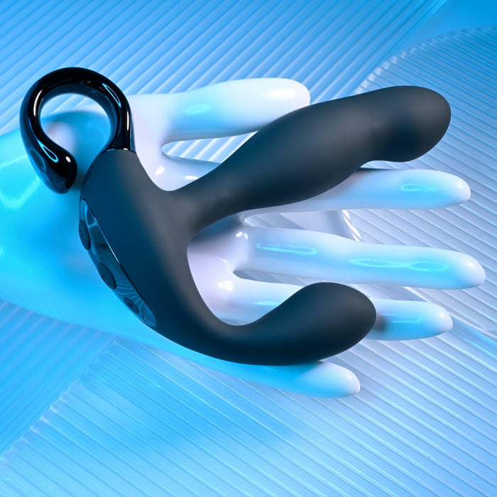 A black Playboy Come Hither Motion Vibrating Prostate Massager by Evolved Novelties, featuring a dual-ended silicone design with a loop handle and capable of 10 vibration speeds, rests on an artificial white hand. The background showcases a blue-tinted, textured surface.