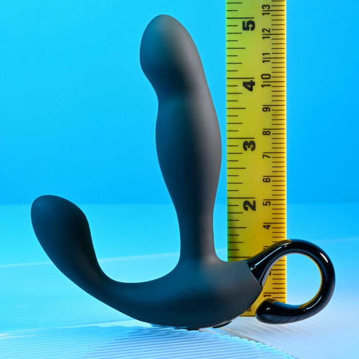 The Playboy Come Hither Motion Vibrating Prostate Massager by Evolved Novelties, a black curved device with two bulbous ends and a looped handle, stands upright next to a vertical yellow ruler, measuring approximately 5.5 inches. Featuring 10 vibration speeds and USB rechargeable capabilities, the background is a smooth surface with a gradient blue color.