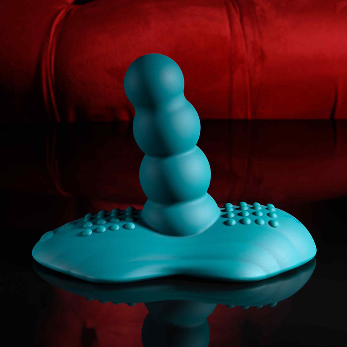 The Playboy Have a Seat Remote Control Grinding Vibrator Pad, an adult toy in blue body-safe silicone with a beaded design and textured base, stands upright on a black surface against a plush red background.
