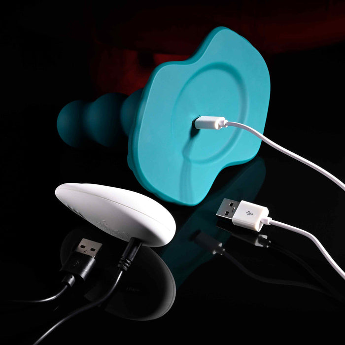 The Playboy Have a Seat Remote Control Grinding Vibrator Pad, featuring a teal silicone design and white oval remote, with USB charging cables, is displayed on a reflective black surface against a dark background.