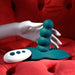 A white mannequin hand holds the Playboy Have a Seat Remote Control Grinding Vibrator Pad, featuring a teal beaded design on a textured suction cup base. A matching white remote with black buttons rests nearby on the red tufted background.
