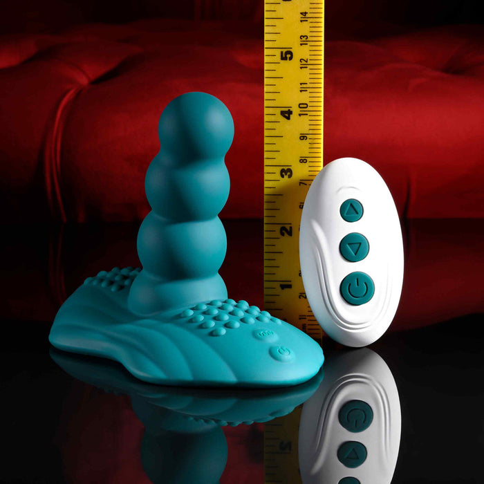 The Playboy Have a Seat Remote Control Grinding Vibrator Pad, made of body-safe teal silicone with a textured base, is shown beside a white remote and yellow measuring tape on a red cushioned background.