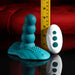 The Playboy Have a Seat Remote Control Grinding Vibrator Pad, made of body-safe teal silicone with a textured base, is shown beside a white remote and yellow measuring tape on a red cushioned background.
