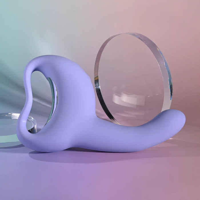 The Playboy Nice to Meet You Powerful Thrusting Vibrator with Handle, a sleek lavender silicone toy with a curved design, rests on a soft pastel surface beside clear, circular acrylic shapes.