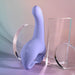 The Playboy Nice to Meet You Powerful Thrusting Vibrator with Handle, a lavender silicone toy featuring a curved, bulbed end and ergonomic base, stands upright among clear geometric acrylic shapes on a pastel background.