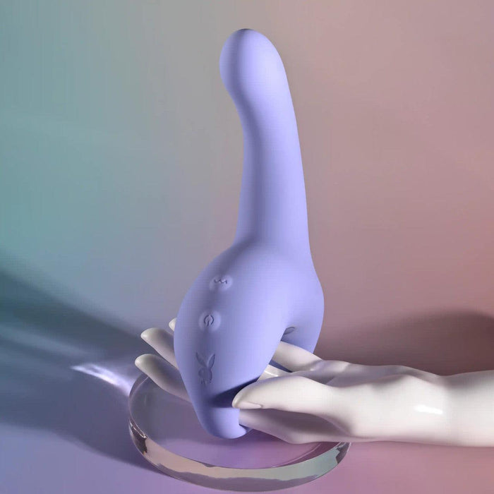 A pale mannequin hand holds the Playboy Nice to Meet You Powerful Thrusting Vibrator with Handle—lavender silicone, curved ergonomic design, and control buttons—displayed on glass against a soft pastel gradient background.