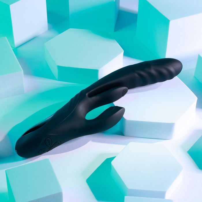 The Playboy Rapid Rabbit Thrusting Vibrator with Flapping Shaft, a sleek and ergonomic black clitoral stimulator, rests on a modern geometric backdrop of white and cyan hexagons, creating a striking contrast with its smooth design.