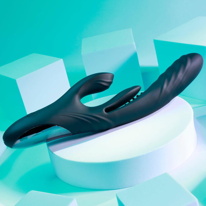 The Playboy Rapid Rabbit Thrusting Vibrator with Flapping Shaft, a black ergonomic adult toy for g-spot stimulation, rests on a white circular platform. Its sleek design stands out against the turquoise gradient background, surrounded by white geometric shapes.