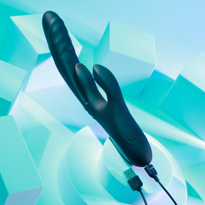 The Playboy Rapid Rabbit Thrusting Vibrator with Flapping Shaft, in black, designed for g-spot stimulation and equipped with a USB charging cable, is displayed on a geometric backdrop featuring cubes and cylinders under teal lighting.